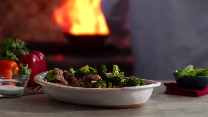 Lock down shot of beef with broccoli with chef flambeing wok in the background.
