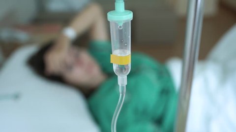 A sick woman lays in a hospital bed behind an IV drip