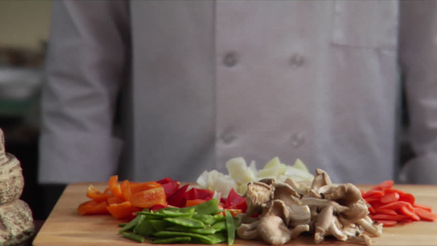 Lock down shot of chef gathering chopped onions and tossing them into a wok, Asian vegetables on a cutting board in the forefront. Royalty-Free Stock Footage #2541572