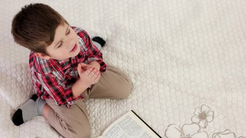 little kid praying on knees, christian kid reading holy bible, small boy with eyes closed saying prayer at home, strong belief in heart, child sitting on knees holding hands together and pray to god
