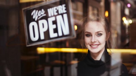 Attractive young blonde woman owner turns sign from close to open and shares a friendly, bright smile to camera. Inside the shop. Being friendly. Successful morning, having a break.