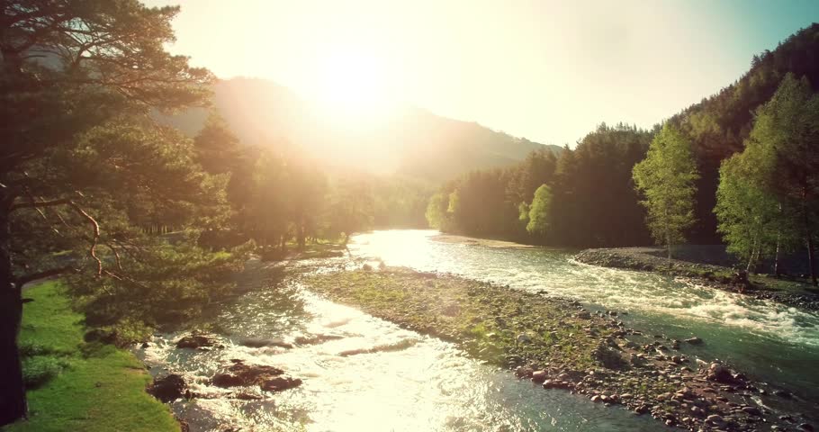 4k (UHD) aerial view. Low flight over fresh cold mountain river at sunny summer morning. Green trees and sun rays.