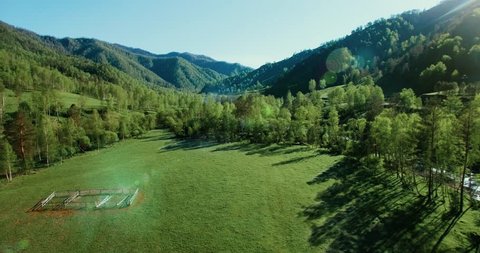 UHD 4K aerial view. Low flight over fresh cold mountain road, river and meadow at sunny summer morning. Green trees and sun rays on horisont.