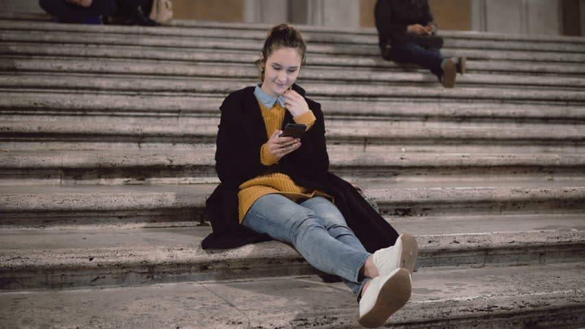 Attractive young student sitting on the ladder and using smartphone. Girl chatting with touchscreen technology