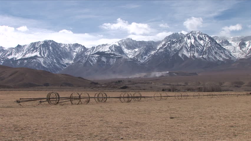 A large wide open field in front of The Sierra Nevada Mountains, which are snow capped and have clouds rolling over them during the day