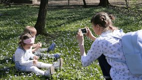 Young Mother Shoot Phone Video Of Two Little Children Sitting On A Green Grass In Park