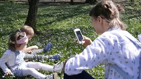 Young Mother Shoot Phone Video Of Two Little Children Sitting On A Green Grass In Park