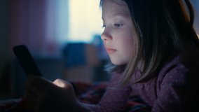 Cute Little Girl in Her Room at Night, Lies on a Bed Uses Smartphone. Screen Illuminates Her Face. Shot on RED EPIC-W 8K Helium Cinema Camera.