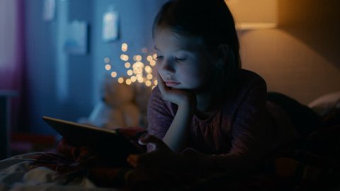 Cute Little Girl in Her Room at Night, Lies on a Bed with Tablet Computer. Her Night Lamp Turned On. Shot on RED EPIC-W 8K Helium Cinema Camera.