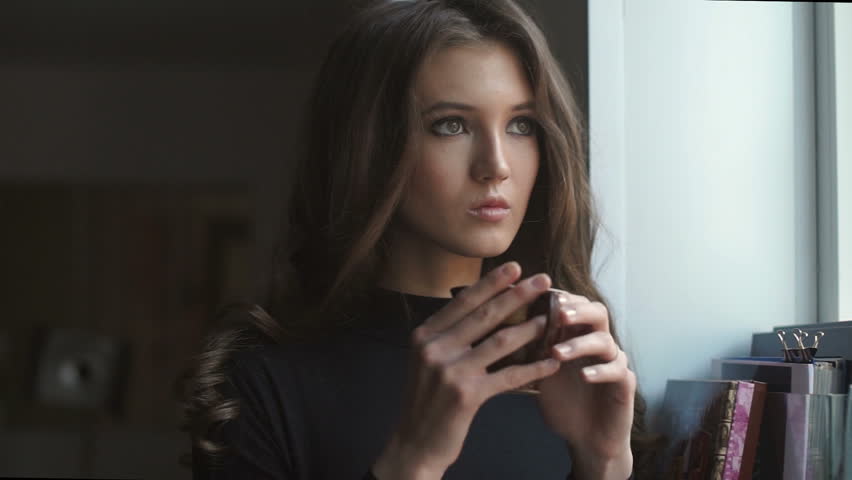 Beautiful young girl looks through the window and drinks coffe.