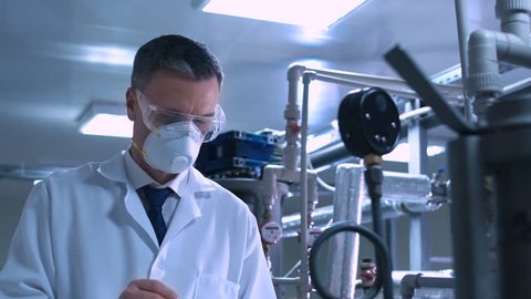 Side view video of male scientist in white lab coat, goggles and mask standing next to productional equipment checking indicators and writing in journal. Middle shot 4K