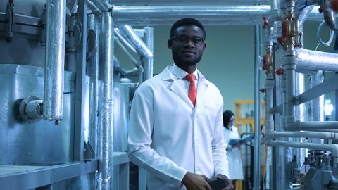 An African smiling scientist looking at camera standing in a plant. Horizontal indoors shot.