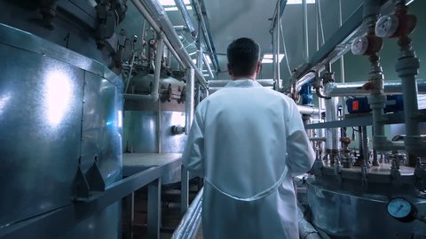 Rear view video of unrecognizable male scientist in white uniform walking through lad with metal tubes and engineering equipment aside