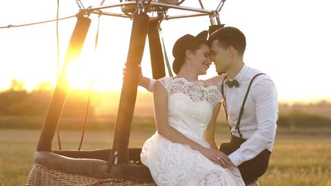 The vintage dressed couple are holding hands and laughing while sitting on the moving air-balloon located in the sunny field during the sunset.