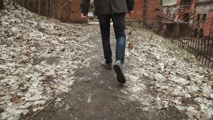 A young man With a beard, in a gray coat walks through the park in the spring or autumn trees yellow leaves, next to an abandoned German fort, brick walls, an old iron fence