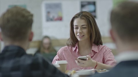 Tracking shot of young beautiful businesswoman using smart phone while sitting at table with male colleagues and discussing something in the office at coffee break