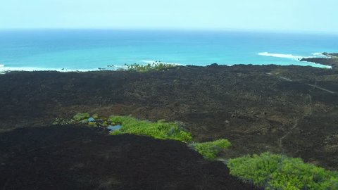 Aerial shot starting from a small green oasis and pond, pulling back to reveal stark lava rock fields and the bright blue Pacific Ocean and hazy Big Island sky, Kona, Hawaii.