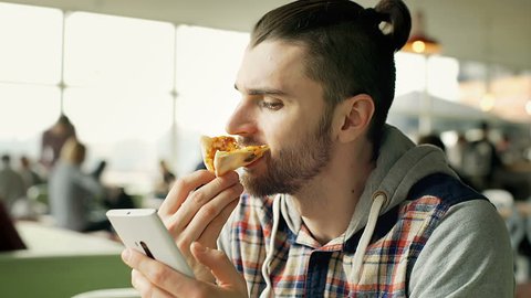 Handsome man eating tasty pizza and browsing internet on smartphone, steadycam shot
