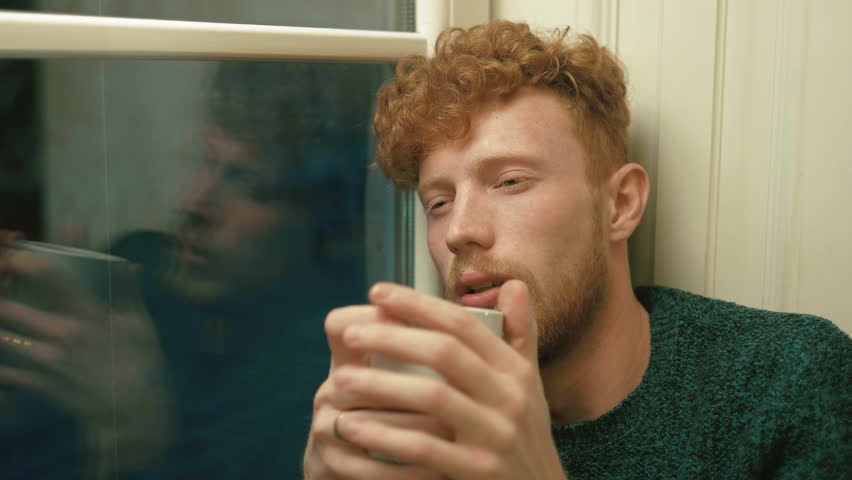 The ill man with golden curly hair holding the white cup, drinking hot tea, coughing and sitting close to the window. Side close-up portrait