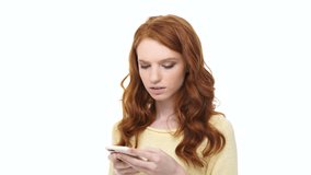 Young displeased annoyed woman with long red hair typing message on smartphone over white background