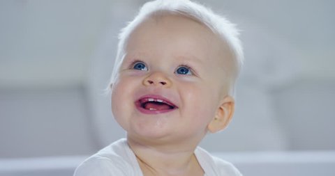 A baby, a boy with large blue eyes and light-colored hair, sits and smiles on a snow-white blanket, looks at her mother, on a white background. Concept: children, kids, baby, babies, a new generation.