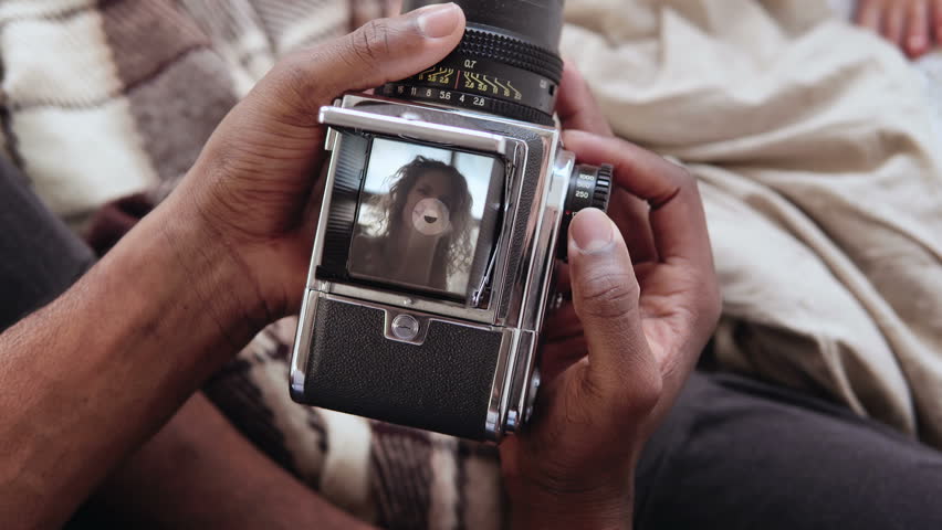 Close-up view of african man holding old photocamera, taking photo portrait beautiful woman. Multiracial couple on bed.