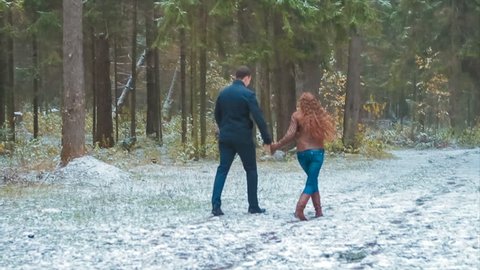 Walk Two Lovers of Young People in Snowy Park on Road Among Tall Pines. Red-Haired Girl in Brown Jacket and Blue Jeans Clings to Hand of Her Tall Guy in Black. They go Opposite Direction From Camera.