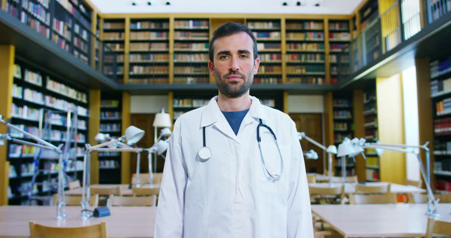 A young and beautiful doctor in a library smiling happy and holding books after doing a search and after studying. Concept: educational, portrait, library, and medical care and welfare