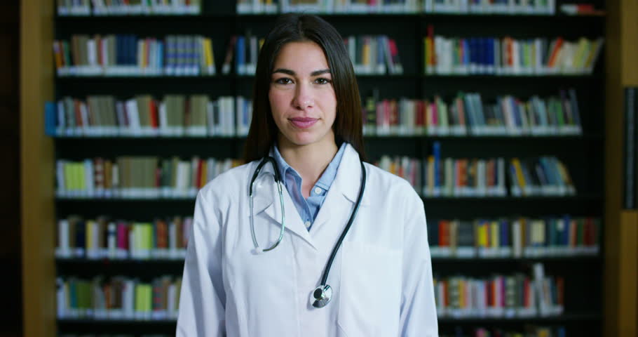 A young and beautiful doctor in a library smiling happy and holding books after doing a search and after studying. Concept: educational, portrait, library, and medical care and welfare