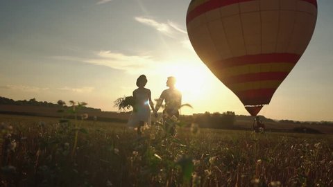The romantic front view of the wintage dressed newlyweds holding hands and walking in the field during the sunset at the background of the colourful air-balloon.