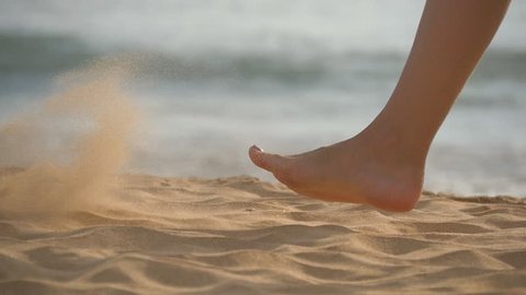 Close up of female feet walking on golden sand at the beach with ocean waves at background. Legs of young woman stepping at sand. Barefoot girl at the sea shore. Summer vacation holiday. Slow motion