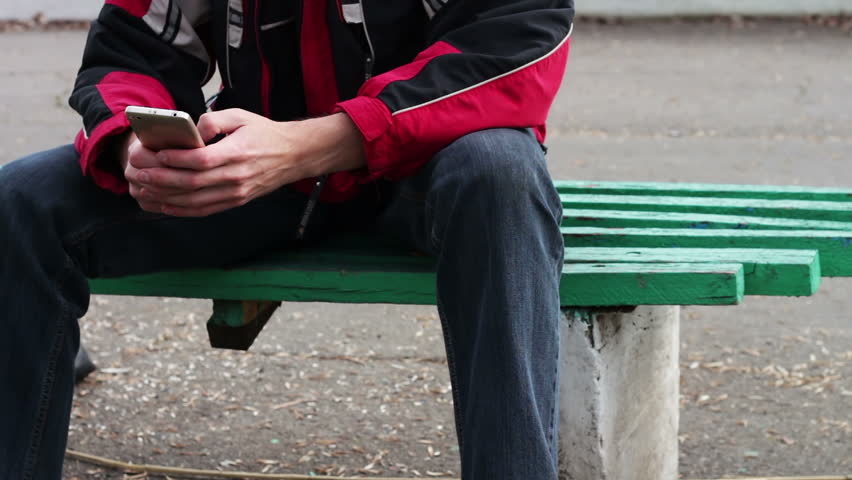 A lonely man sits in a park on a bench waiting for someone and typing something on the smartphone