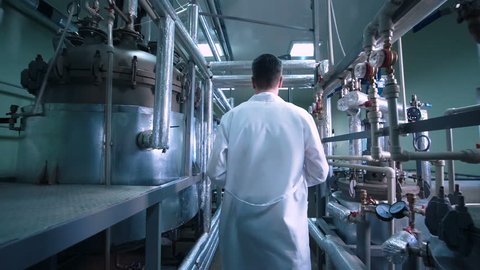 Rear view video of unrecognizable male scientist in white uniform walking through lad with metal tubes and engineering equipment aside