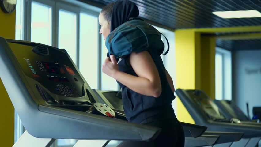 Pretty sporty woman is engaged on a treadmill in a fitness club, changing the intensity of the run. A woman is running with a bag on her shoulders to increase the load on her legs.
