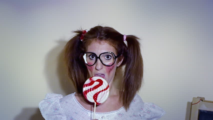 A woman dressed up like a little girl, with nerdy glasses, licking a lollipop while looking at the camera - zoom in, she says some nonsense words. Funny comedic shot.
