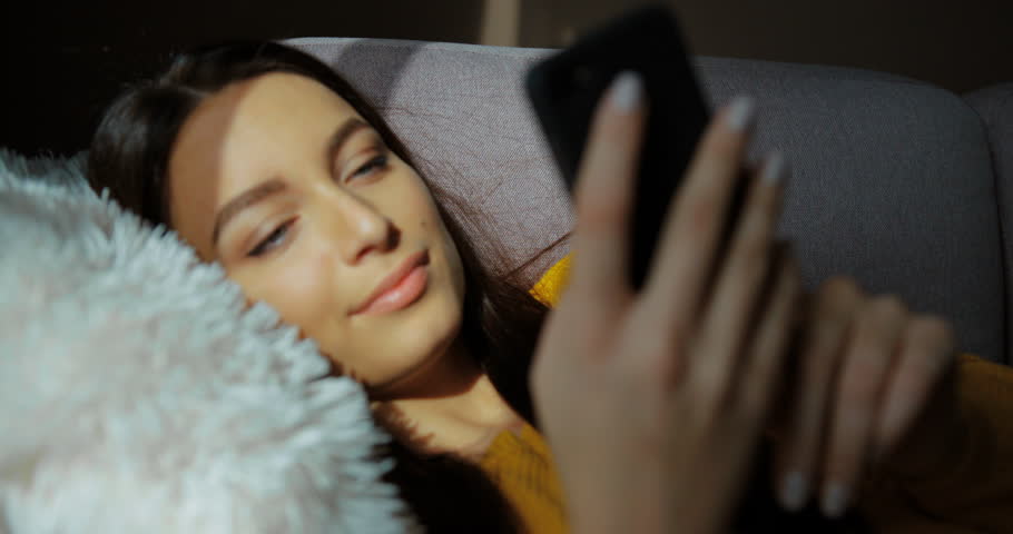 Attractive young woman typing on her smart phone while lying on the sofa at home. Smilling woman using smart phone, chatting, web surfing