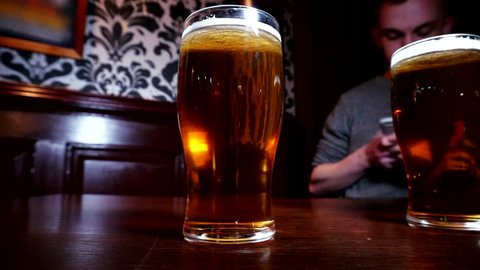 Time lapse of two pints of beer toast with two young men drinking in a pub / bar at night. Alcohol and socializing, heavy binge drinking fast. Pints of alcoholic lager drinks. Dark scene