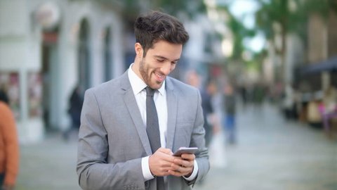 Handsome and young businessman using a cell phone in a crowded street. He is checking mails, chats or the news online. Technology.