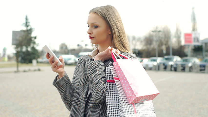 Annoyed girl checking time on smartphone and waiting for someone with shopping bags
