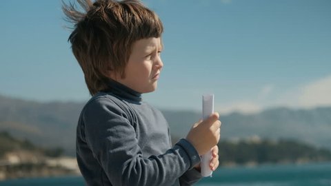 boy folded piece of paper looking into the distance like trumpet to mountains. Slow motion picture of child standing on lake holding hands with paper in cage, like telescope, closed right eye, looks