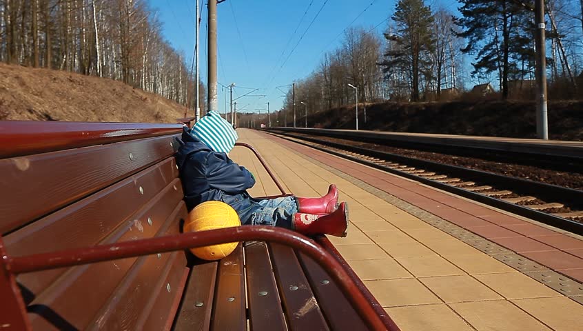 video boy in a jacket and hat sits on a bench in a railway station in the spring with a yellow football.