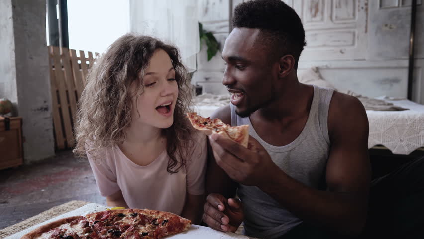 Happy multiethnic couple in pajamas eating fast food. Hungry woman waits the meal, man feeds her a slice of pizza. | Shutterstock HD Video #25802858
