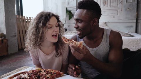 Happy multiethnic couple in pajamas eating fast food. Hungry woman waits the meal, man feeds her a slice of pizza.