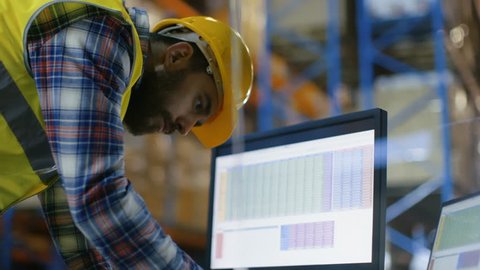 Male Inspector Wearing Hard Hat Fills in Spreadsheets on His Personal Computer. He's in Big Warehouse with Rows of Pallet Racks. Shot on RED EPIC-W 8K Helium Cinema Camera. 