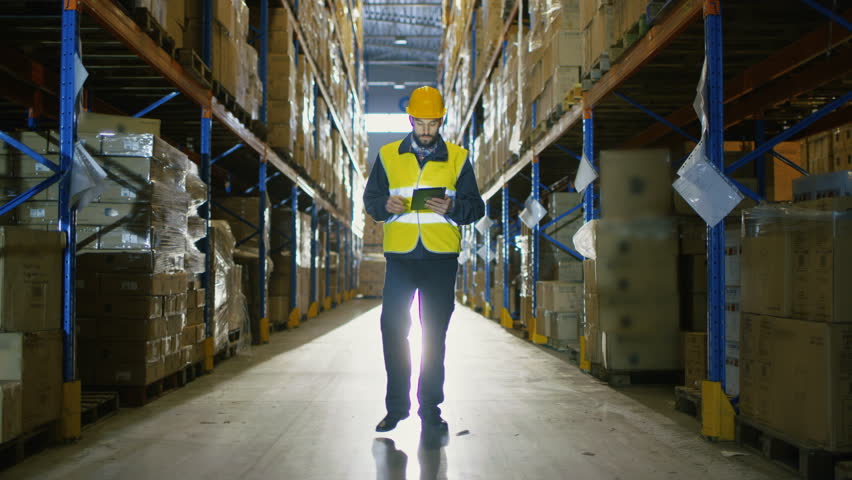 Auditor Wearing Hard Hat with Tablet Computer Counts Merchandise in Warehouse. He Walks Through Rows of Storage Racks with Merchandise.  Royalty-Free Stock Footage #25809242