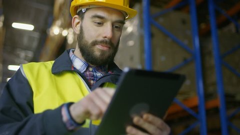 Surveyor Wearing Hard Hat Holds Tablet Computer and Counts Merchandise in Warehouse full of Racks with Boxes on Them. 