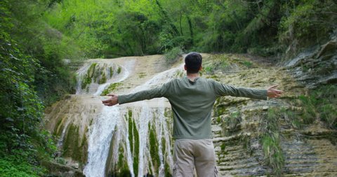 Happy young man, breathes in himself a breath of air to transform the qi, feels free and independent , on the background of a beautiful waterfall and rocks, concept: nature, religion, bio, ecology.