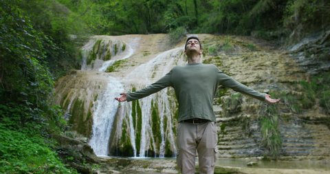 Happy young man, breathes in himself a breath of air to transform the qi, feels free and independent , on the background of a beautiful waterfall and rocks, concept: nature, religion, bio, ecology.