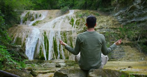 Sitting young man, found a wonderful place for yoga practice, feels free and independent , on the background of a beautiful waterfall and rocks, the concept: nature, religion, bio, ecology, qi.