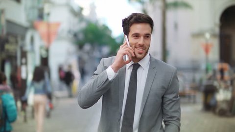 Handsome and young businessman using smartphone in a crowded street. He is talking on the phone and laughing while walking. City.

